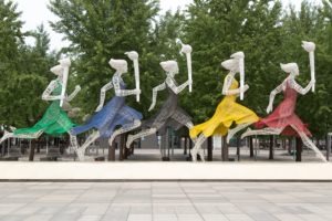 five running women wearing different dresses holding torch statue