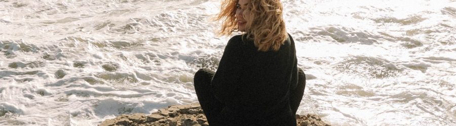 woman in black long sleeve shirt sitting on brown rock near sea during daytime